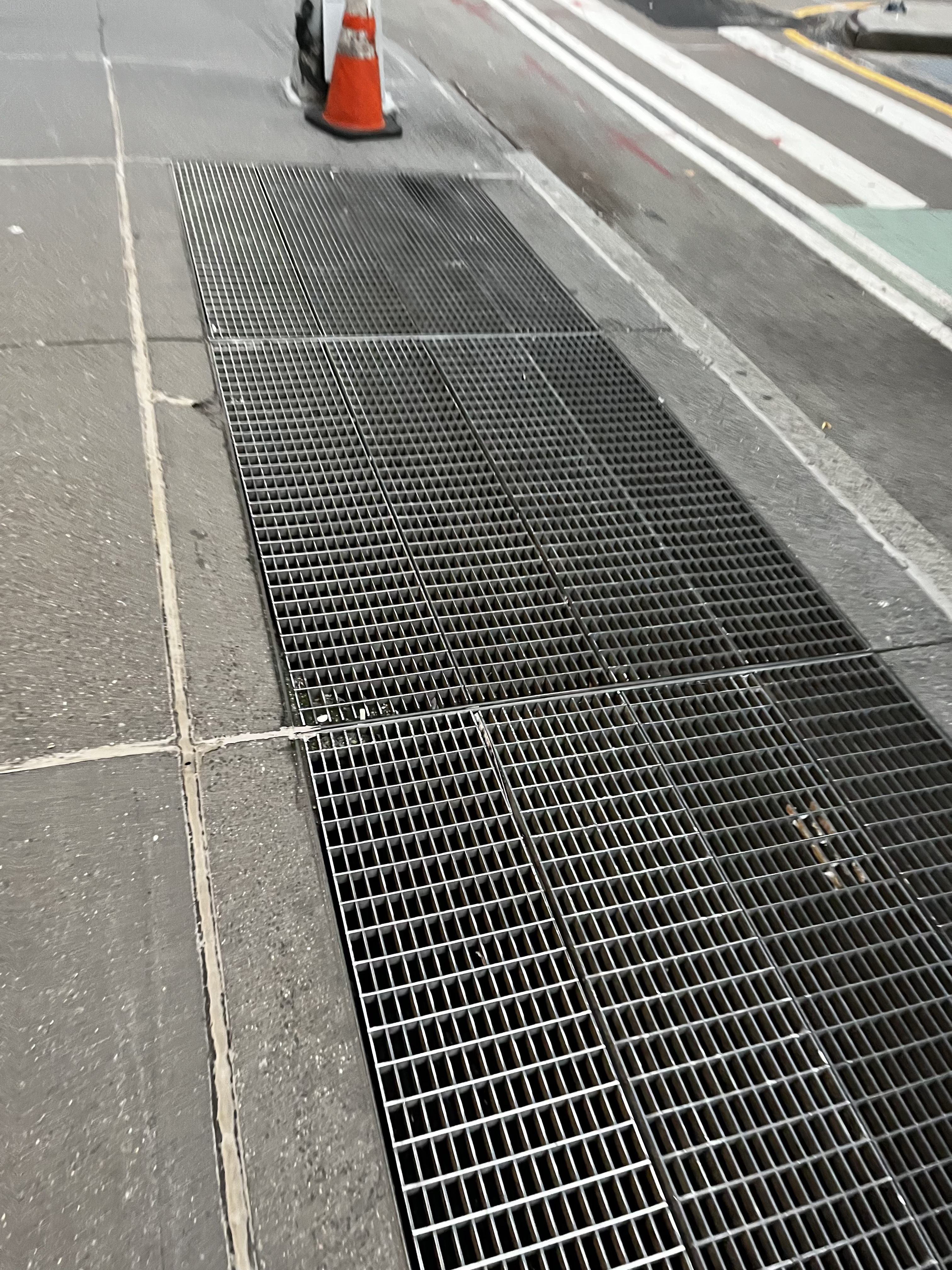 City grate: geometric pattern, safety orange cone nearby.